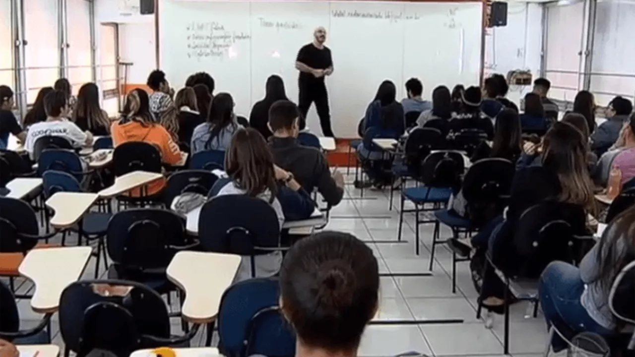 Sala de aula vista do fundo onde alunos prestam a atenção a professor, que leciona na frente da sala